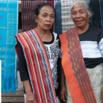 Two older women wearing bright, striped woven sashes stand together in front of hanging textiles at a market stall.