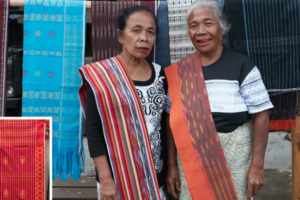Two older women wearing bright, striped woven sashes stand together in front of hanging textiles at a market stall.