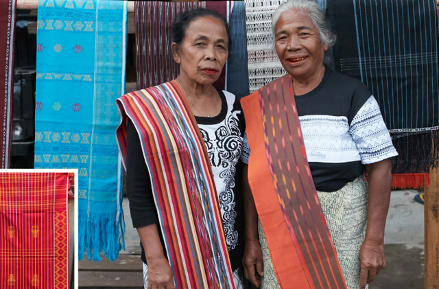Two older women wearing bright, striped woven sashes stand together in front of hanging textiles at a market stall.