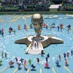 Large golden trophy centerpiece on a raised platform, surrounded by a circular formation of colorful performers on a bright blue arena floor.