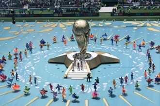 Large golden trophy centerpiece on a raised platform, surrounded by a circular formation of colorful performers on a bright blue arena floor.