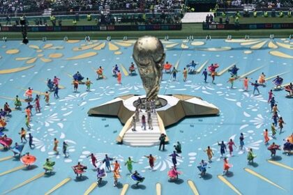 Large golden trophy centerpiece on a raised platform, surrounded by a circular formation of colorful performers on a bright blue arena floor.