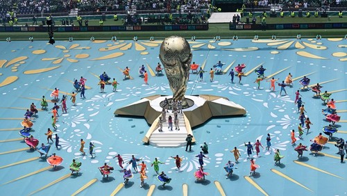 Large golden trophy centerpiece on a raised platform, surrounded by a circular formation of colorful performers on a bright blue arena floor.