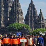 Group of people wearing bright orange shirts posing in front of ancient stone temple towers on a sunny day, greenery in the foreground.