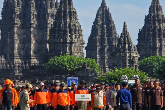 Group of people wearing bright orange shirts posing in front of ancient stone temple towers on a sunny day, greenery in the foreground.