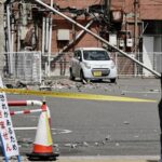 Small white car on a taped-off street after a collapsed utility pole and rubble; a pedestrian passes by.