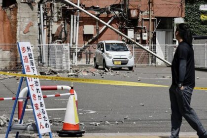Small white car on a taped-off street after a collapsed utility pole and rubble; a pedestrian passes by.