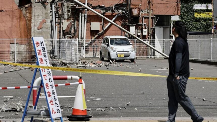 Small white car on a taped-off street after a collapsed utility pole and rubble; a pedestrian passes by.