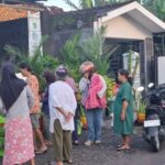 Group of people in hijabs and casual clothing stand near a street market stall with baskets and plants outside a small building; a motorcycle and bicycles are parked nearby.