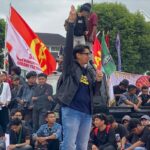 Speaker on a raised platform with a microphone, raising his right hand as crowds and colorful flags loom behind him at a protest.
