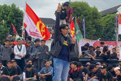 Speaker on a raised platform with a microphone, raising his right hand as crowds and colorful flags loom behind him at a protest.