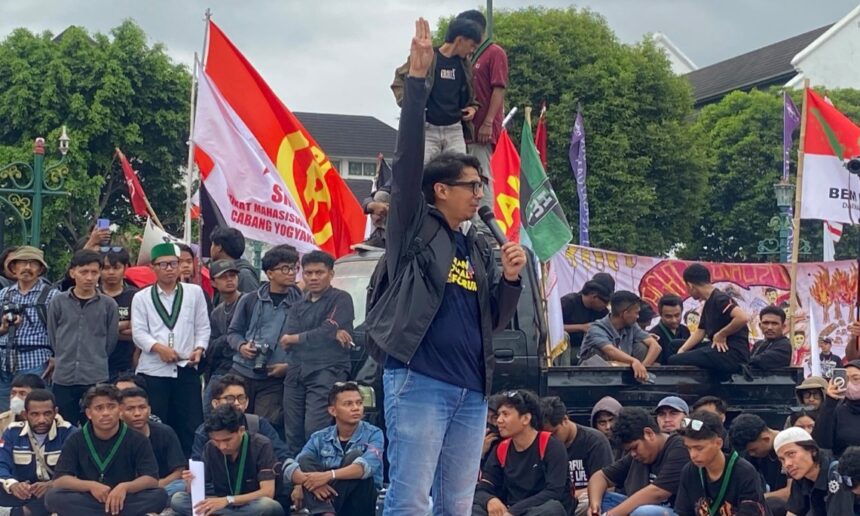 Speaker on a raised platform with a microphone, raising his right hand as crowds and colorful flags loom behind him at a protest.
