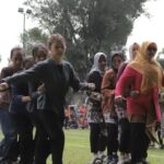 Group of women in hijabs and colorful clothing performing a coordinated line dance on a grassy field at an outdoor event, with spectators in the background.