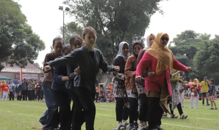 Group of women in hijabs and colorful clothing performing a coordinated line dance on a grassy field at an outdoor event, with spectators in the background.