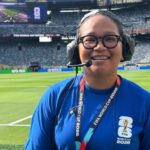 Staff member with headset and glasses smiling on a football pitch, wearing a blue shirt and a FIFA World Cup 2026 lanyard at the stadium union jack background]
