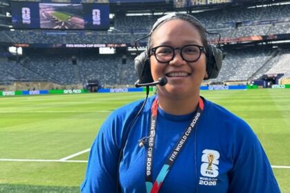 Staff member with headset and glasses smiling on a football pitch, wearing a blue shirt and a FIFA World Cup 2026 lanyard at the stadium union jack background]