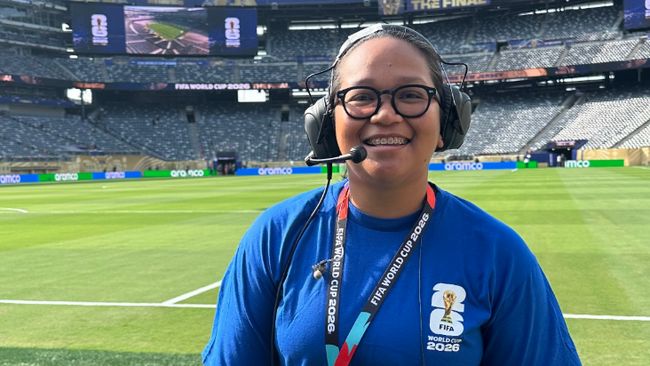 Staff member with headset and glasses smiling on a football pitch, wearing a blue shirt and a FIFA World Cup 2026 lanyard at the stadium union jack background]