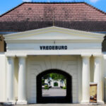 White historic gate labeled VREDEBURG with four columns and a triangular pediment, opening to a tree-lined pedestrian path.