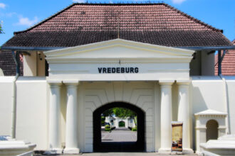 White historic gate labeled VREDEBURG with four columns and a triangular pediment, opening to a tree-lined pedestrian path.