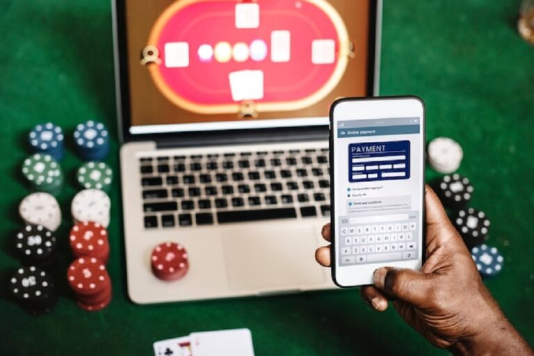 Person holds a smartphone showing an online payment form above a laptop on a green casino table with scattered poker chips