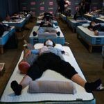 Rows of people lying on air mattresses arranged in neat rows in a dim sleep-lab room, some wearing masks, pillows and blankets visible.