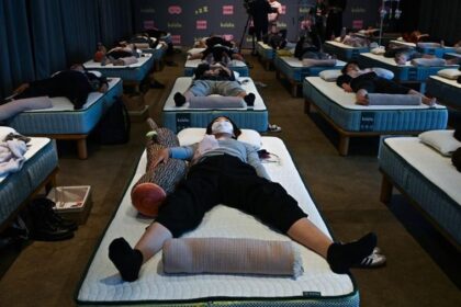 Rows of people lying on air mattresses arranged in neat rows in a dim sleep-lab room, some wearing masks, pillows and blankets visible.
