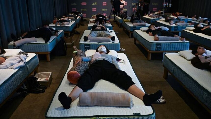 Rows of people lying on air mattresses arranged in neat rows in a dim sleep-lab room, some wearing masks, pillows and blankets visible.