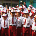 Group of schoolchildren in white shirts, red ties, and white-red caps standing in formation during a school event or assembly.