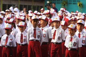 Group of schoolchildren in white shirts, red ties, and white-red caps standing in formation during a school event or assembly.