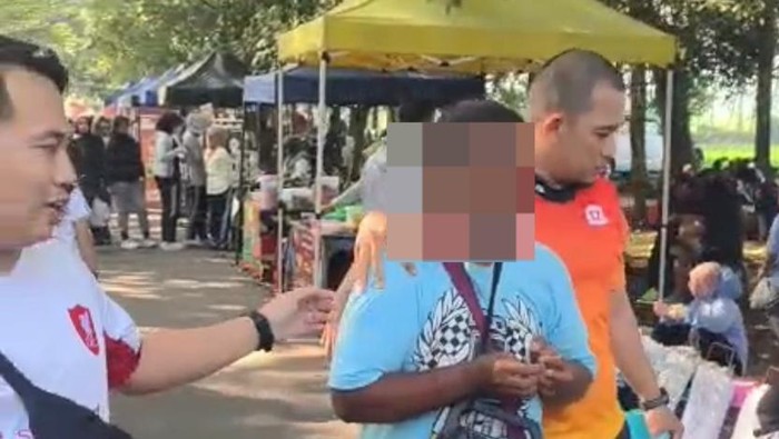 Man in a white shirt hands something to a person with a pixelated face at an outdoor market stall row under tents sunlit scene