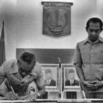 Man signs a document at a ceremonial table as two officials stand nearby in a formal government setting, with portraits on the wall behind him.