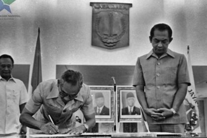 Man signs a document at a ceremonial table as two officials stand nearby in a formal government setting, with portraits on the wall behind him.