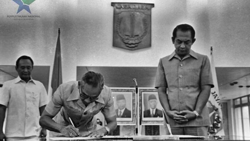Man signs a document at a ceremonial table as two officials stand nearby in a formal government setting, with portraits on the wall behind him.