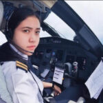 Female pilot in cockpit wearing headset, looking at camera with flight instruments and maps nearby.