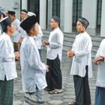 Group of men in white traditional tunics and caps standing in a line outside a building courtyard, chatting.