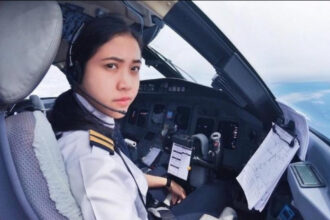 Female pilot in cockpit wearing headset, looking at camera with flight instruments and maps nearby.