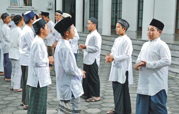 Group of men in white traditional tunics and caps standing in a line outside a building courtyard, chatting.