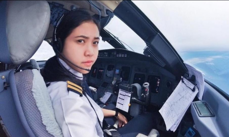 Female pilot in cockpit wearing headset, looking at camera with flight instruments and maps nearby.