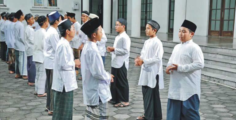 uioo - Mabur.co Group of men in white traditional tunics and caps standing in a line outside a building courtyard, chatting.
