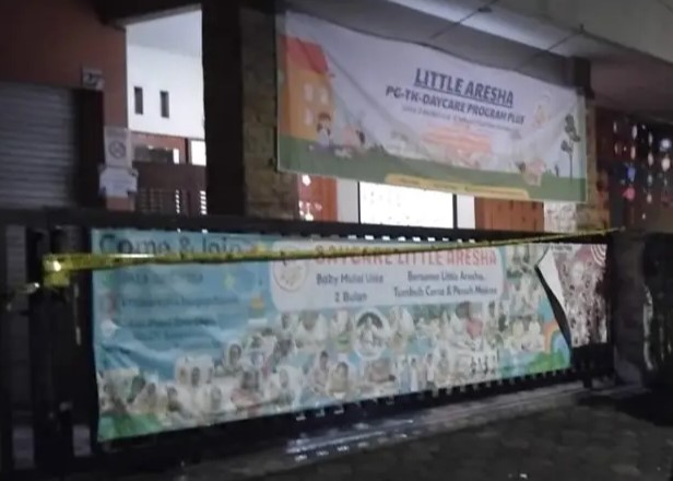 Banner promoting a daycare program 'Little Are_sha' with cartoon children, displayed on a storefront at night; yellow caution tape across railing in front.