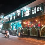 Nighttime street view of a turquoise mosque with arched windows and a colorful sign, a motorbike passing by.
