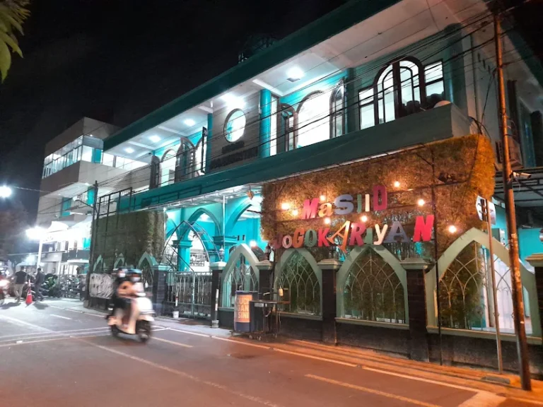 Nighttime street view of a turquoise mosque with arched windows and a colorful sign, a motorbike passing by.