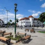 Public square with wooden benches around a planter, ornate green lampposts, and white colonial buildings under a blue sky.