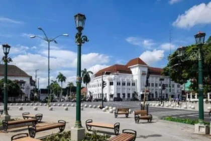 Public square with wooden benches around a planter, ornate green lampposts, and white colonial buildings under a blue sky.