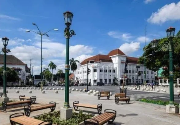 Public square with wooden benches around a planter, ornate green lampposts, and white colonial buildings under a blue sky.