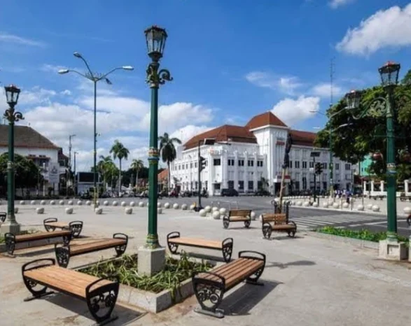 Public square with wooden benches around a planter, ornate green lampposts, and white colonial buildings under a blue sky.