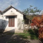 Small white cottage with a cross on the gable, brown wooden shutters, and weathered walls, set in a garden with a red-orange shrub and a tree against a blue sky.