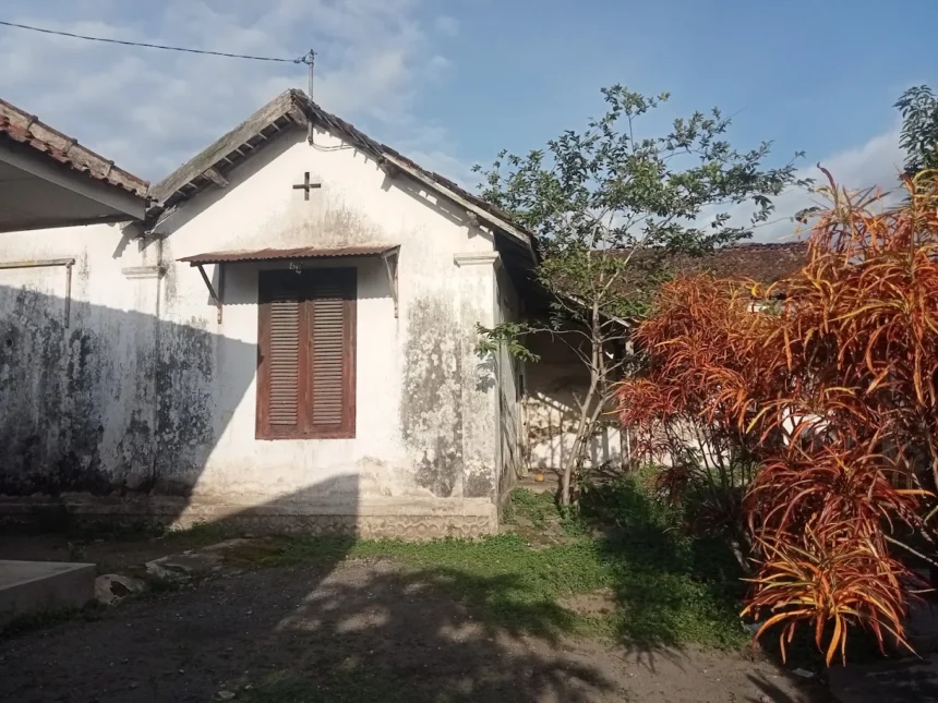 Small white cottage with a cross on the gable, brown wooden shutters, and weathered walls, set in a garden with a red-orange shrub and a tree against a blue sky.