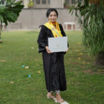Graduate in a black gown with a yellow stole, holding a diploma outdoors on campus.