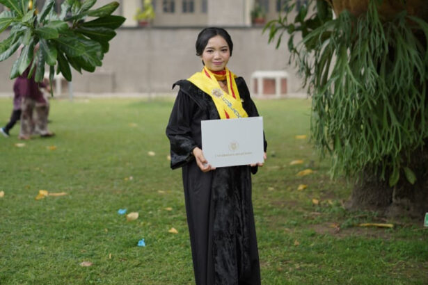 Graduate in a black gown with a yellow stole, holding a diploma outdoors on campus.